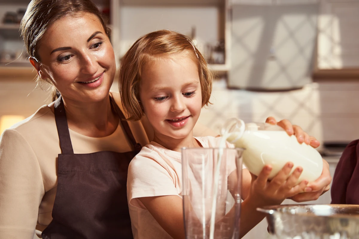 Un enfant qui verse du lait dans un contenant transparent sous le regard de sa maman en tablier