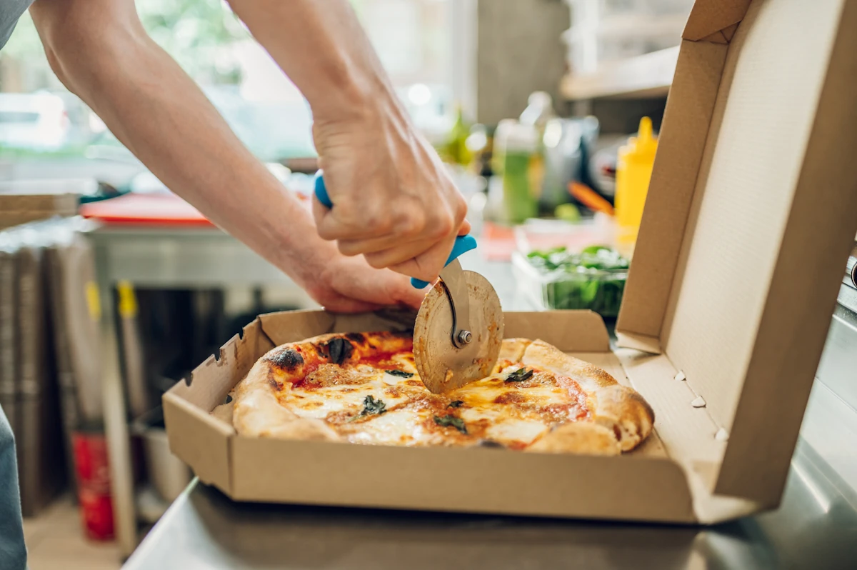Une personne qui coupe une pizza ronde dans une boîte carrée sur une table