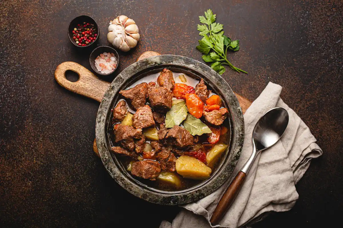 Un bol de pot au feu de viande avec des léhumes posé sur une plaque en bois à coté de quelques ingrédients et une cuillère
